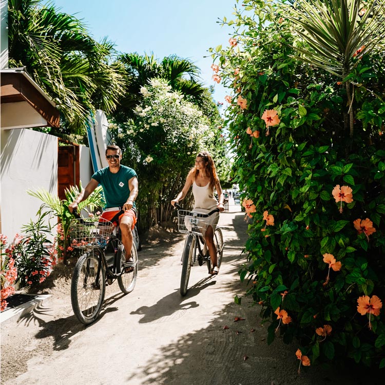 Leon und Sammy fahren auf dem Fahrrad durch die Straßen von Caye Caulker an einem sonnigen Tag neben schönen Blumen in Belize